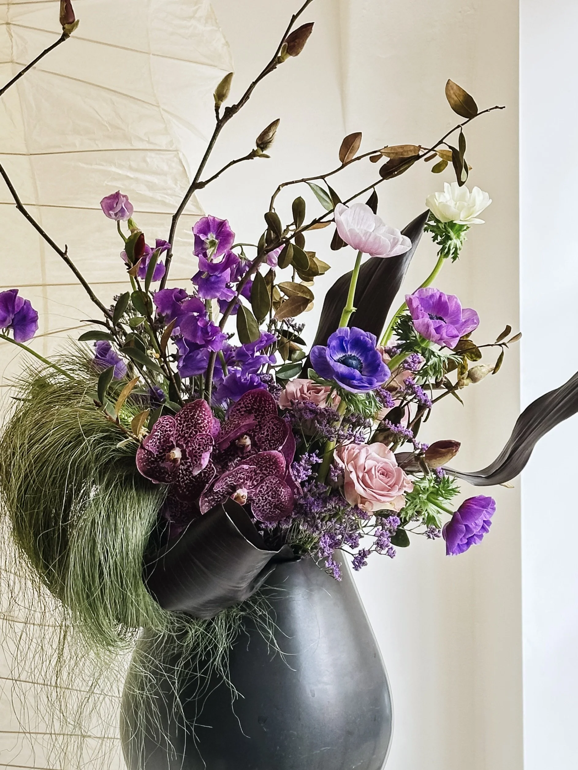 Sculptural floral arrangement featuring orchids, anemones, roses, and dramatic foliage in a dark vessel.