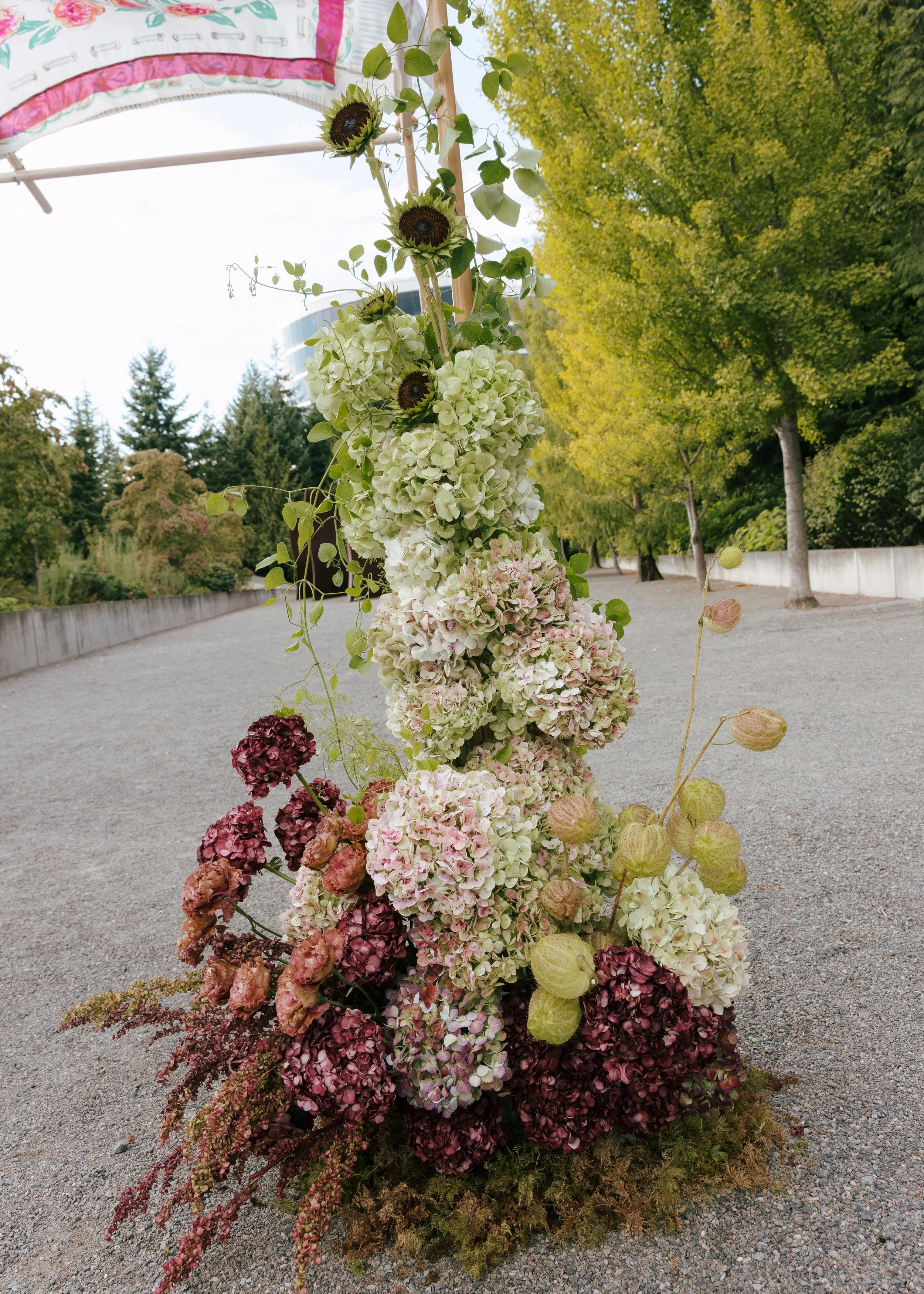 Sculptural wedding ceremony floral installation at Olympic Sculpture Park featuring cascading hydrangeas, sunflowers, and textural seasonal blooms in soft green, blush, and deep burgundy. Floral design by Butterfly’s Rest. Photography by Steph Estrad