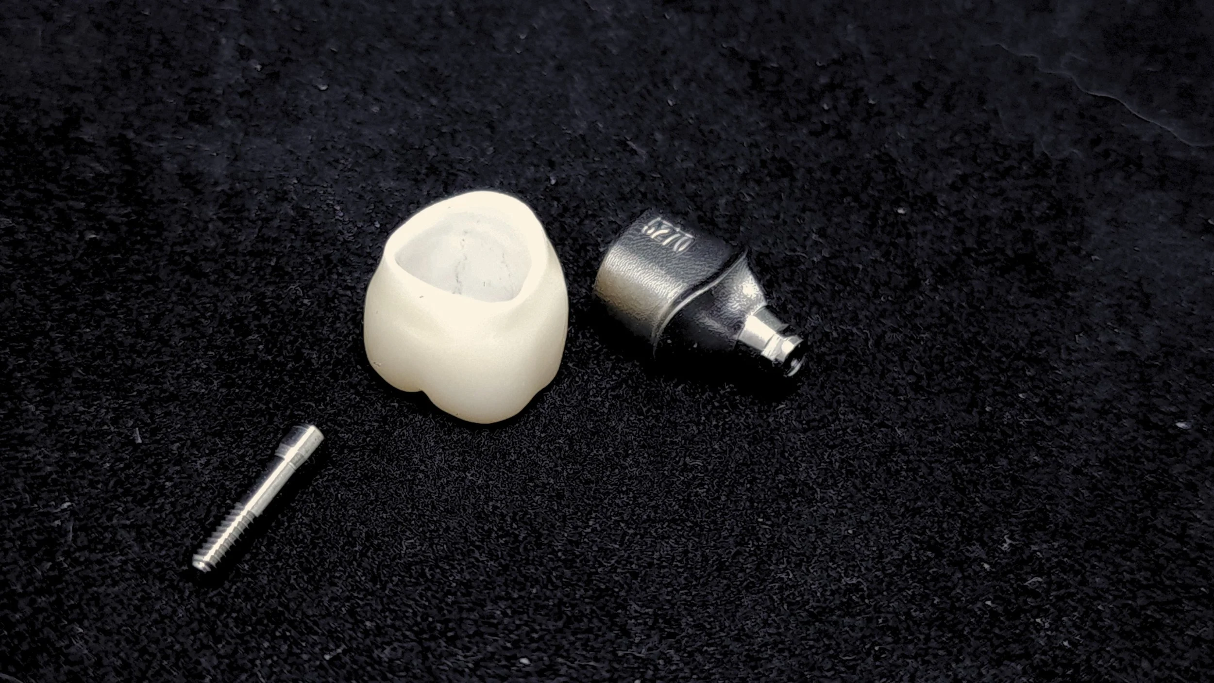Close-up of a small silver object, a white ceramic or plastic piece, and a metal rod on a black textured surface.