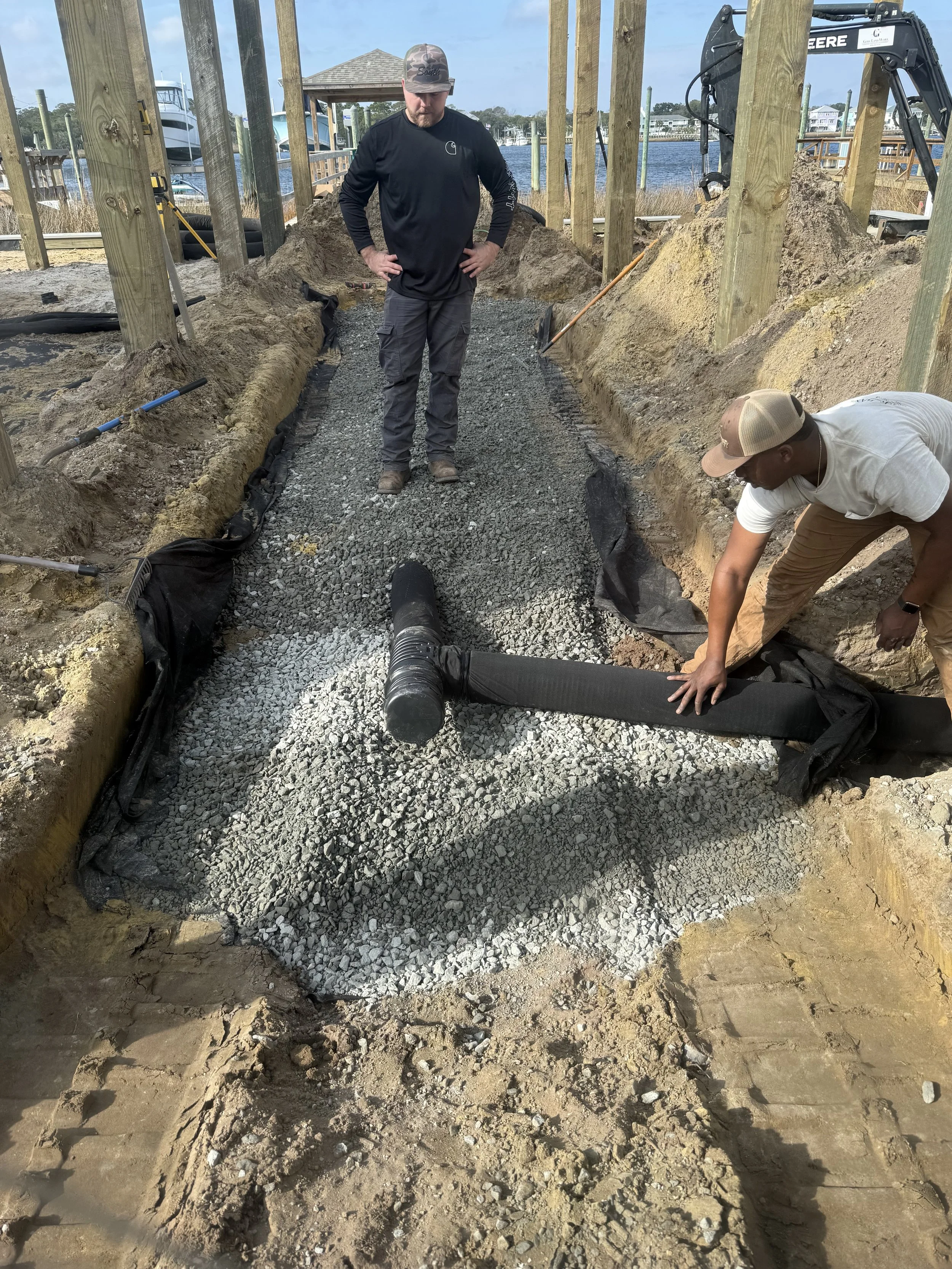 Two men working on a construction project at a waterfront site, laying gravel and drainage pipes with a boat and water in the background.