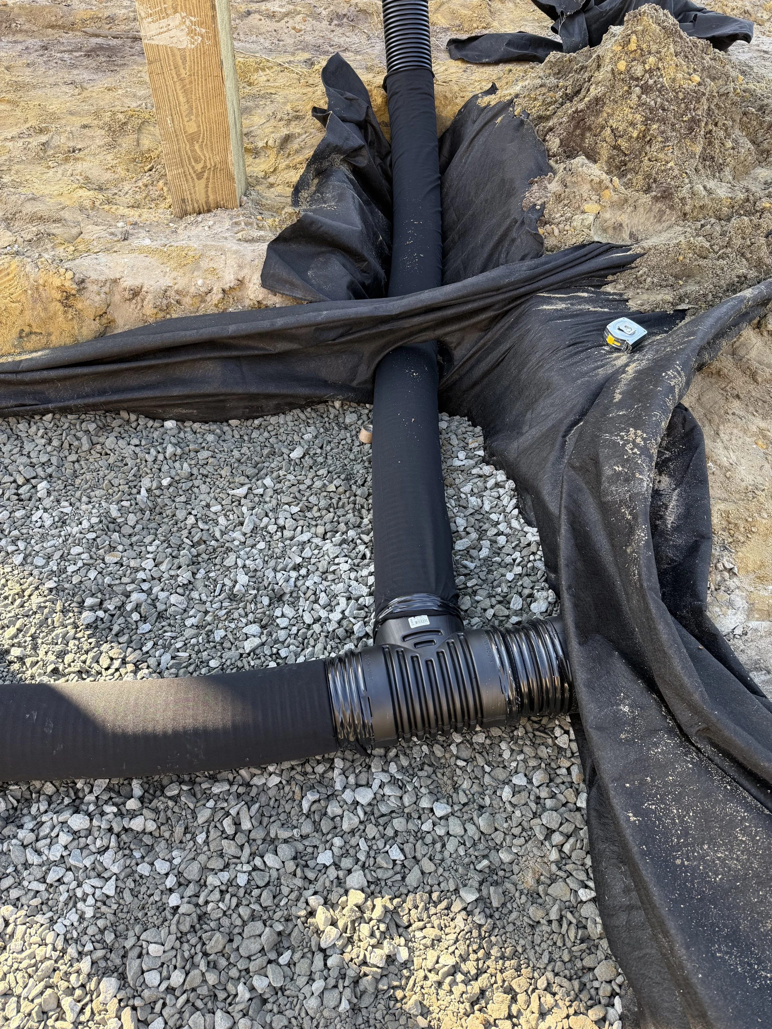 Construction site with black corrugated pipes, drainage pipes, gravel, black fabric, and wooden support post.