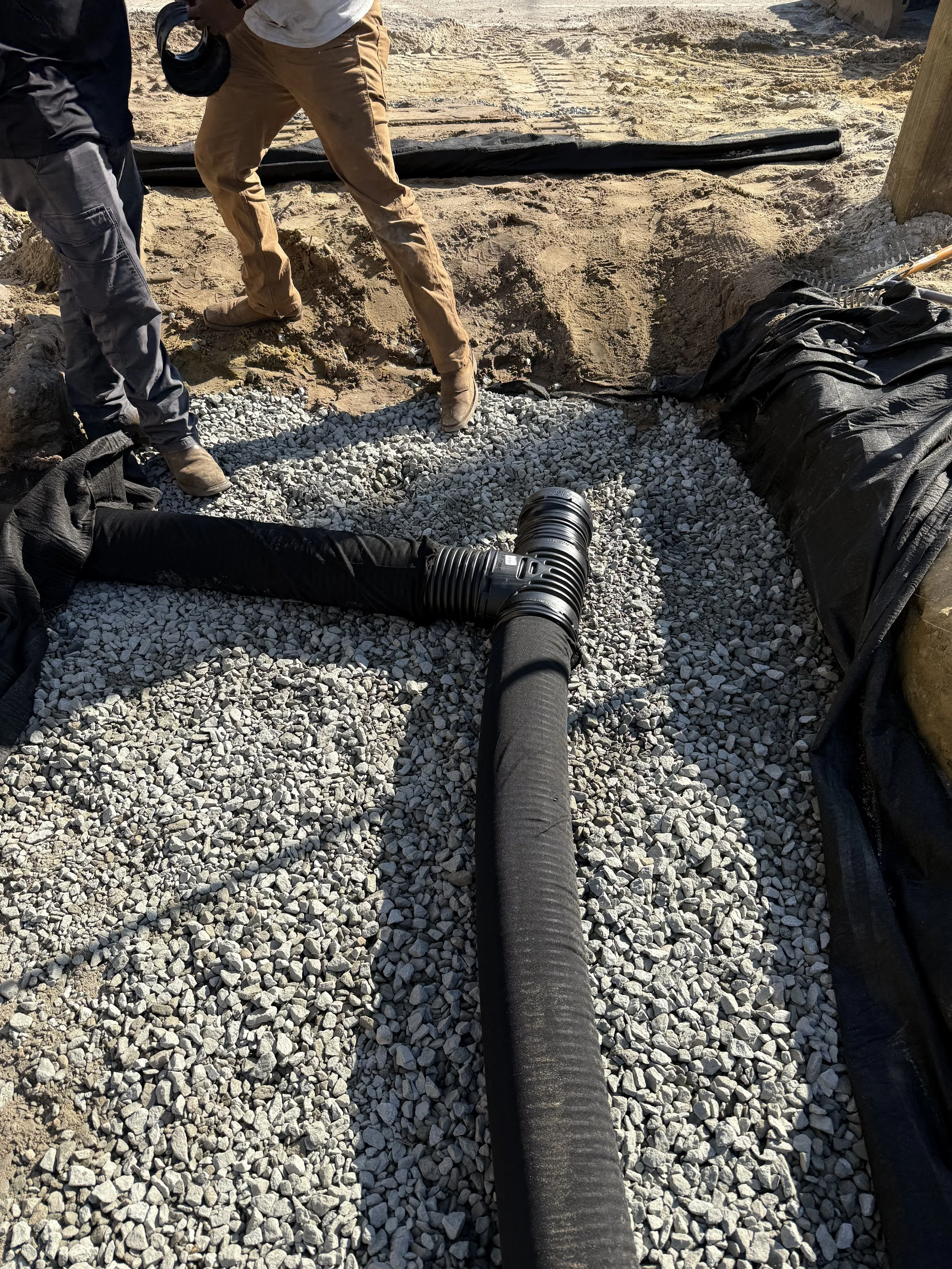 Construction workers are installing a large black drainage pipe into a gravel bed at a construction site, with dirt and wooden posts nearby.