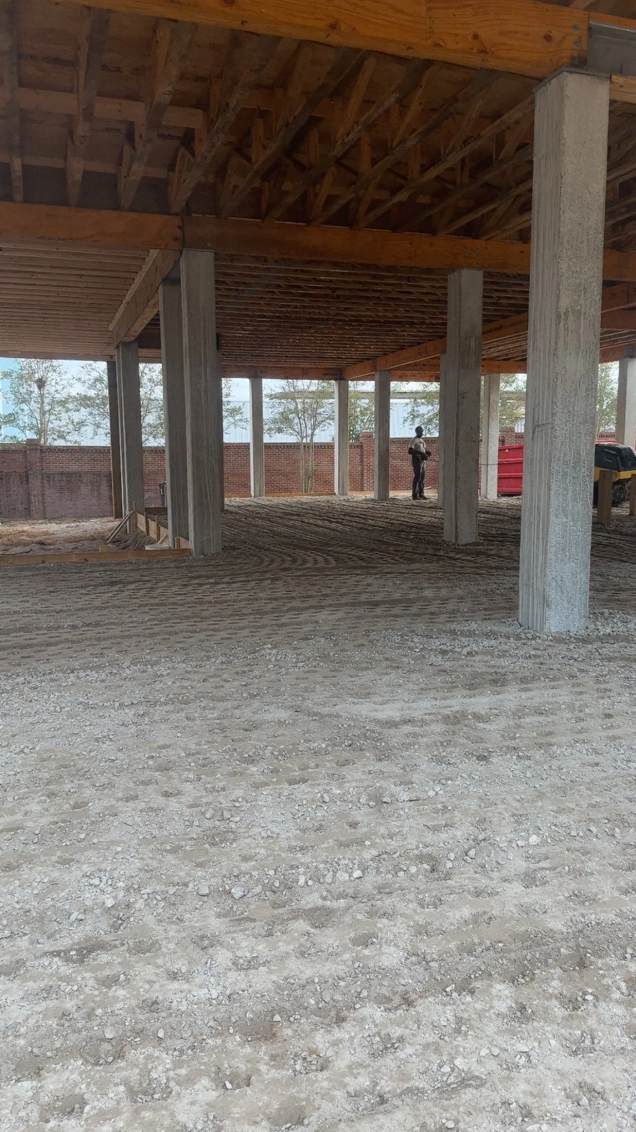 Construction site with wooden beams and concrete columns, dirt ground, and partially built structure, with worker in background.