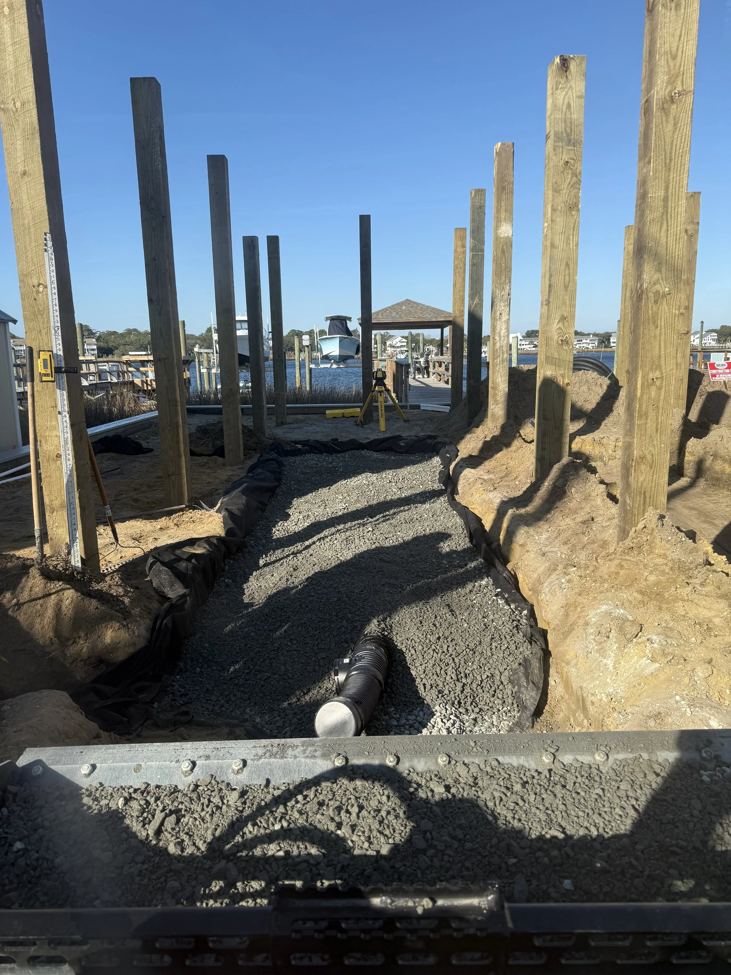 Construction site with wooden posts and gravel pathway near a body of water with boats, umbrellas, and a gazebo under a clear blue sky.