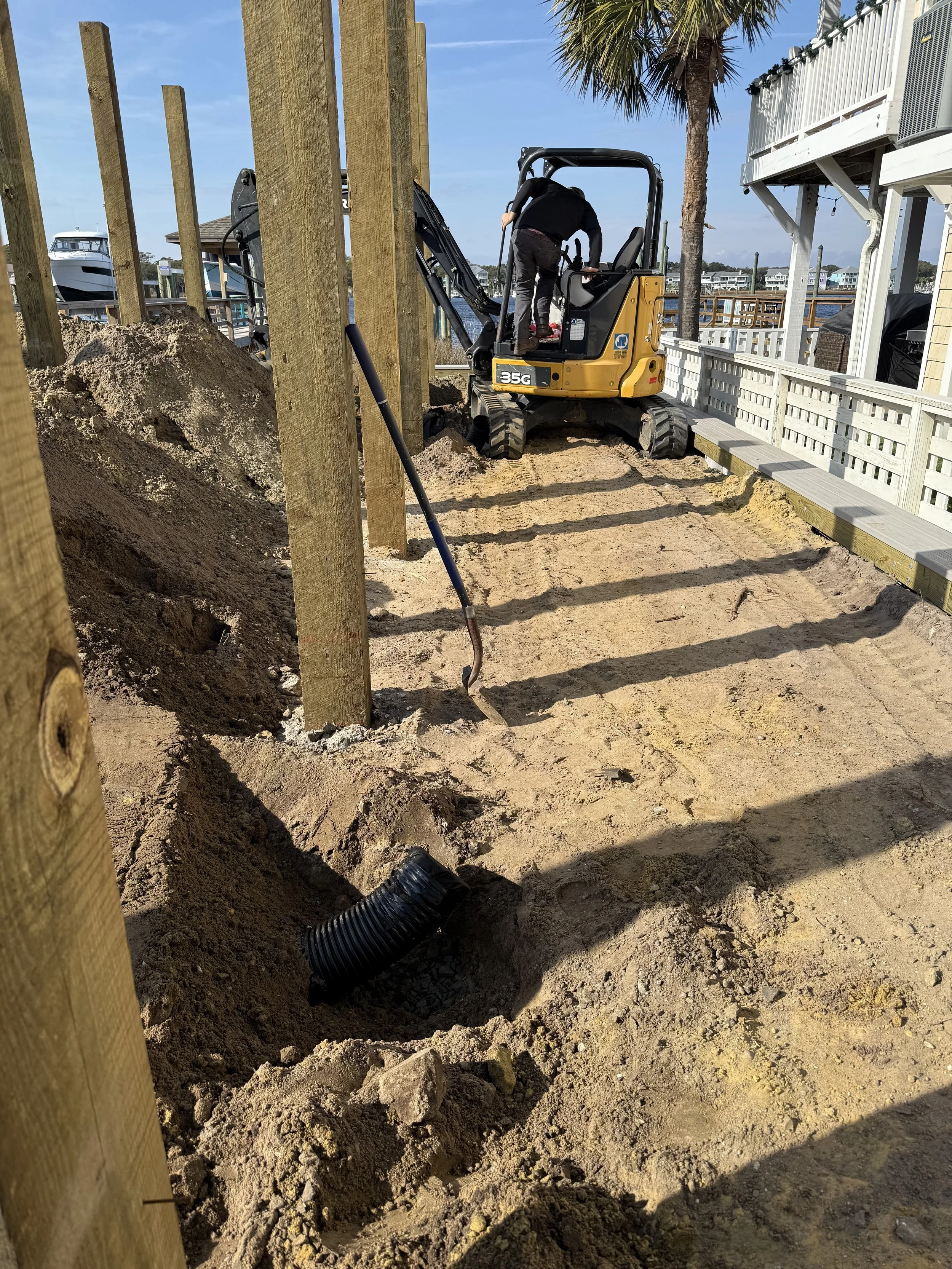 A construction site featuring a small excavator digging in sandy soil, with wooden fencing and a palm tree in the background, near a waterfront with boats.