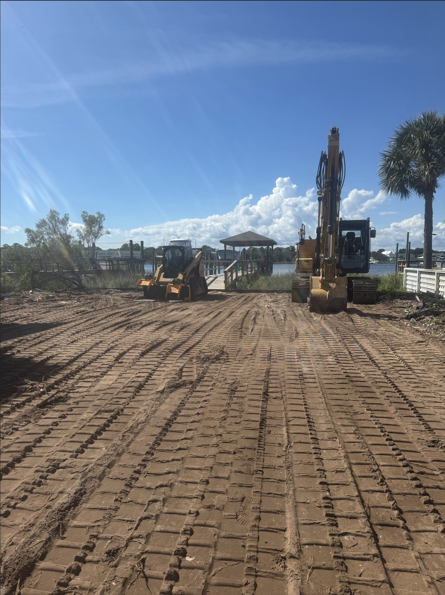 Construction site with dirt road, two yellow excavators, a wooden pier, and a body of water in the background. There are trees, a palm, and a blue sky with clouds.
