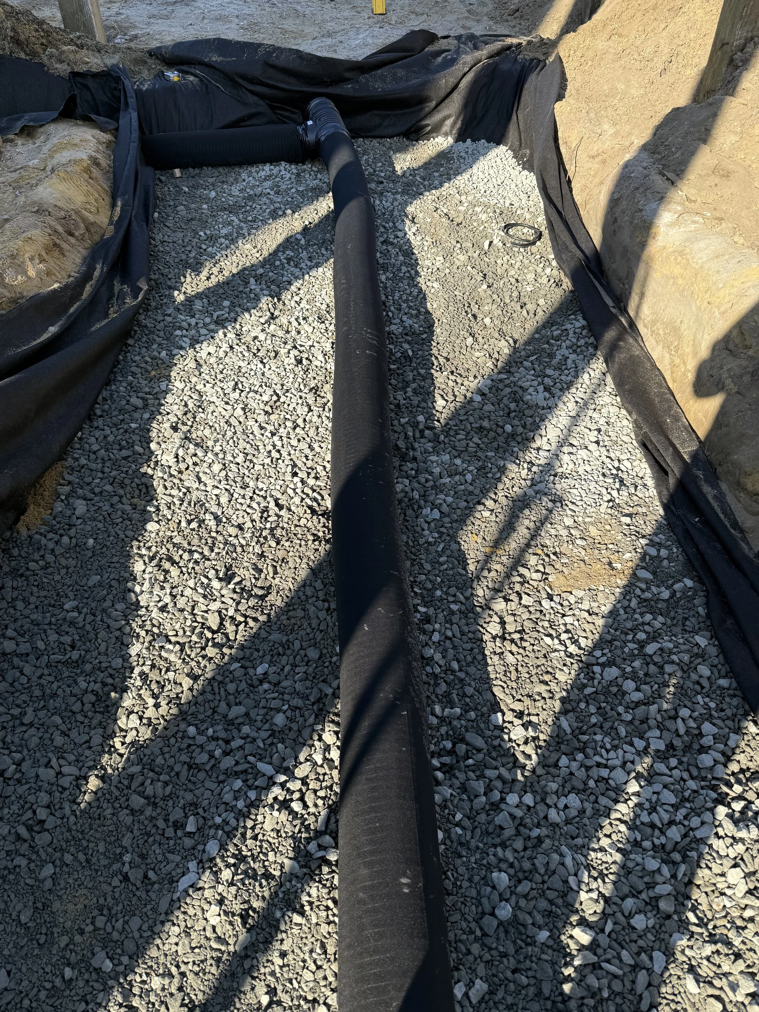 Construction site with black drainage pipe laying across gravel, surrounded by dirt and black geotextile fabric.