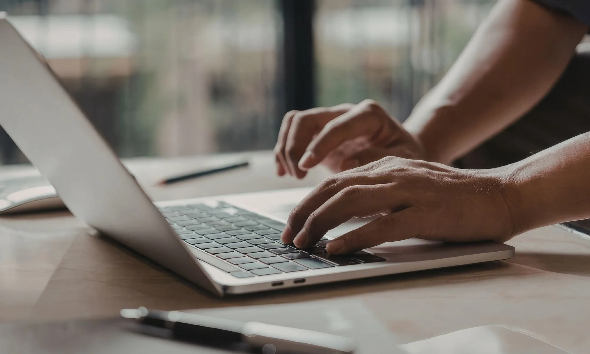 Close-up of hands typing on a silver laptop keyboard placed on a wooden desk with a blurred background.