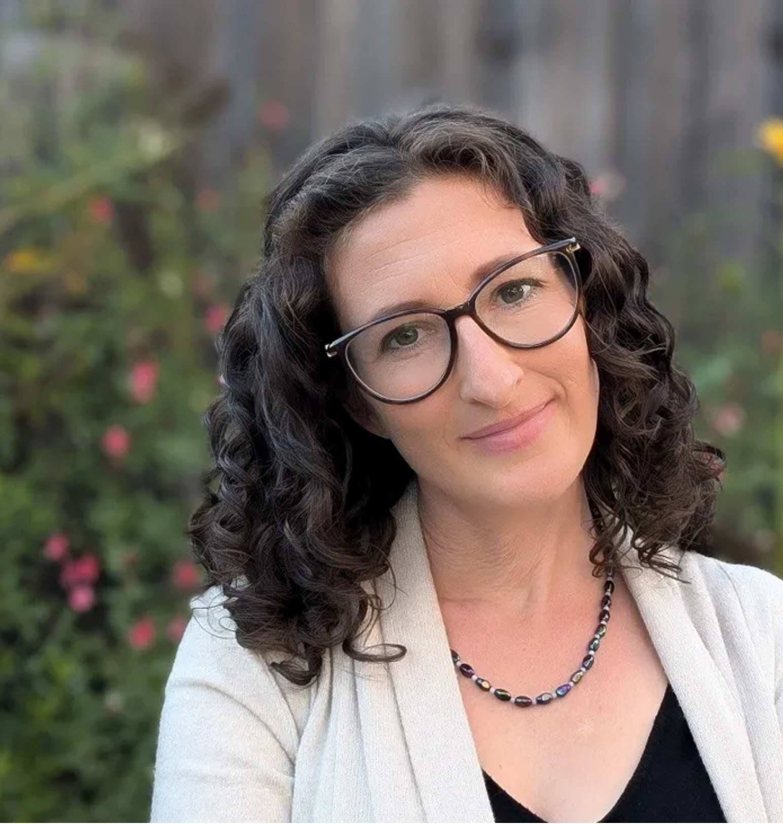 A woman with curly dark hair and glasses smiling outdoors with a blurred garden background.