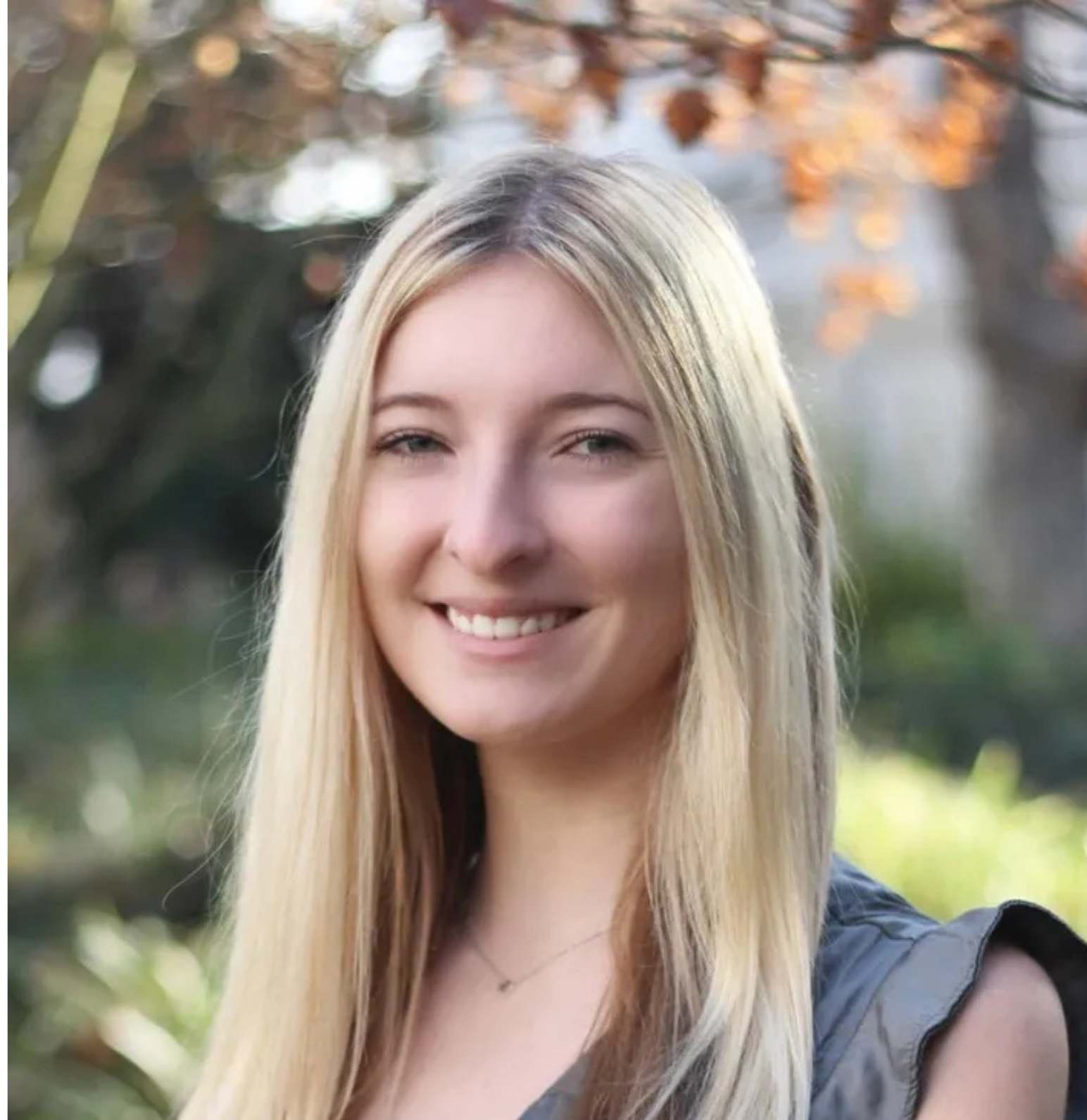 A young woman with long blonde hair smiling outdoors, with autumn leaves and blurred greenery in the background.
