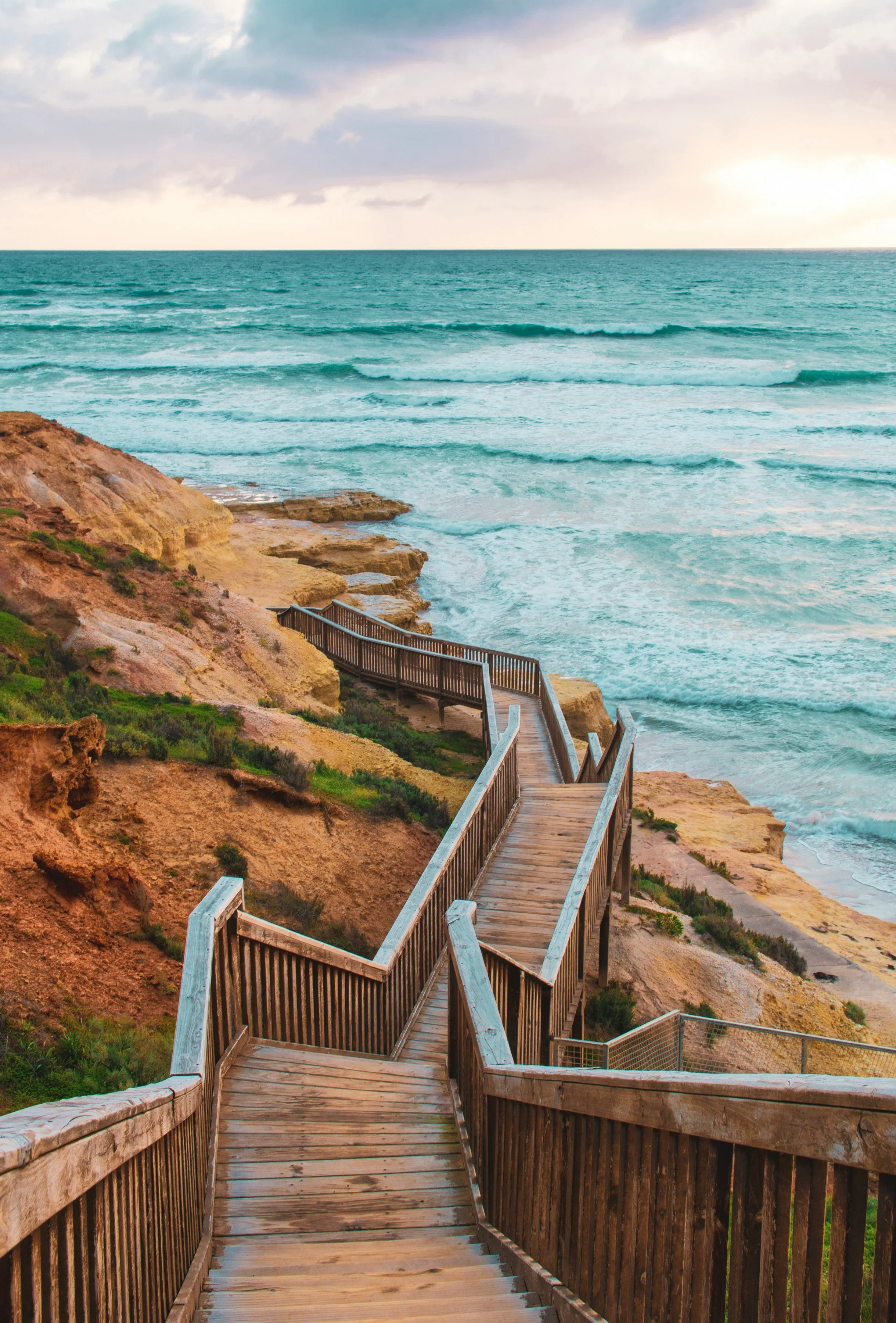 Wooden staircase leading down to the beach with rocky cliffs and ocean waves under a cloudy sky.