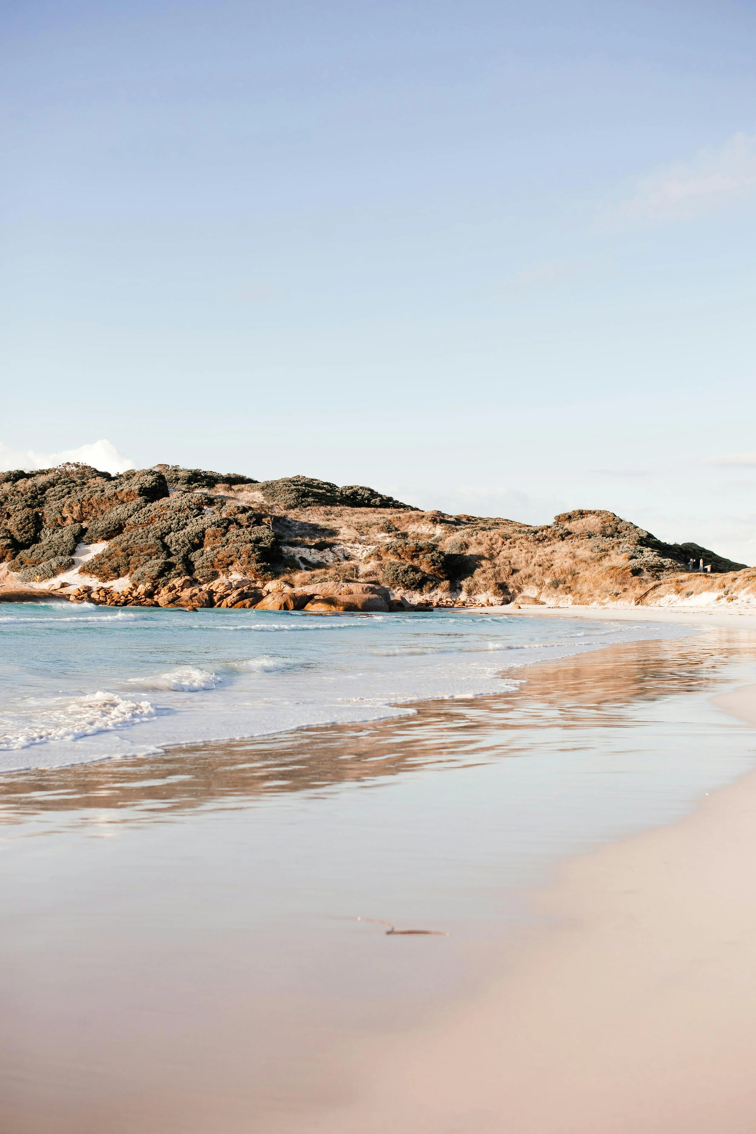 A peaceful beach with clear blue water, gentle waves, sandy shore, and rocky dunes in the background under a partly cloudy sky.