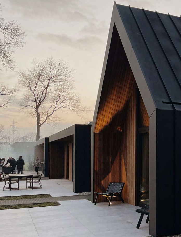 Modern house with black and wood exterior, patio with chairs, and leafless trees in the background during dusk.