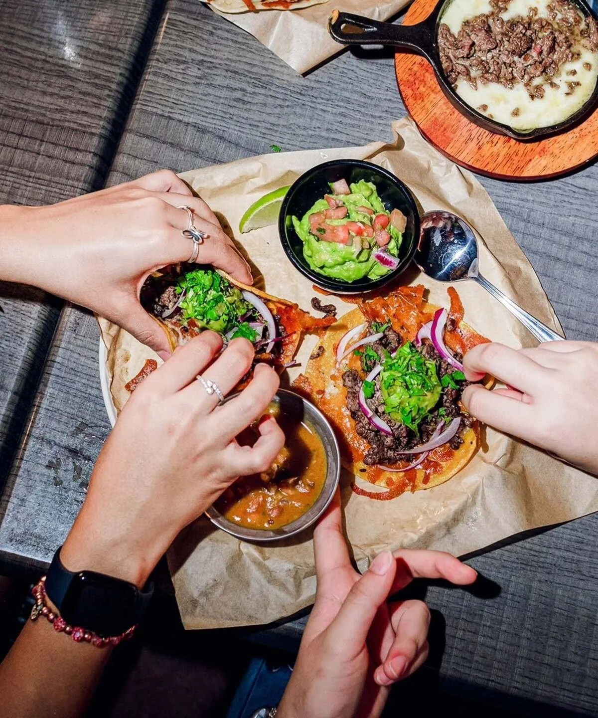 People sharing a meal of tacos with toppings, guacamole, and salsas on a wooden table.