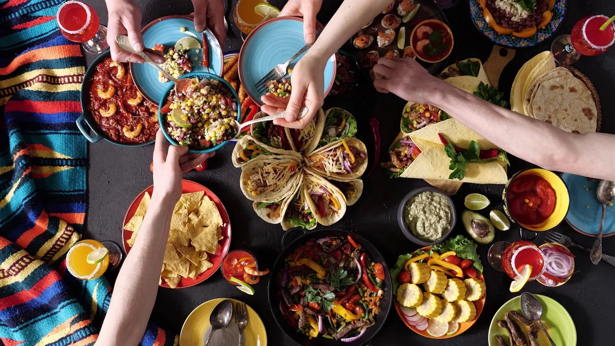 Top-down view of a lively Mexican feast with various dishes including tacos, chips, salsas, salads, and beverages, with hands reaching for food and colorful tableware and tablecloths.