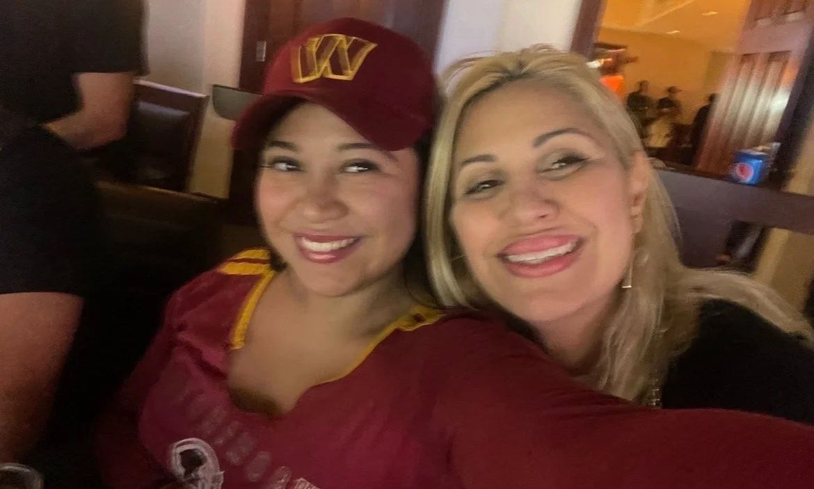 Two women smiling for a selfie in a restaurant. One woman has blonde hair and the other is wearing a maroon baseball cap and jersey.