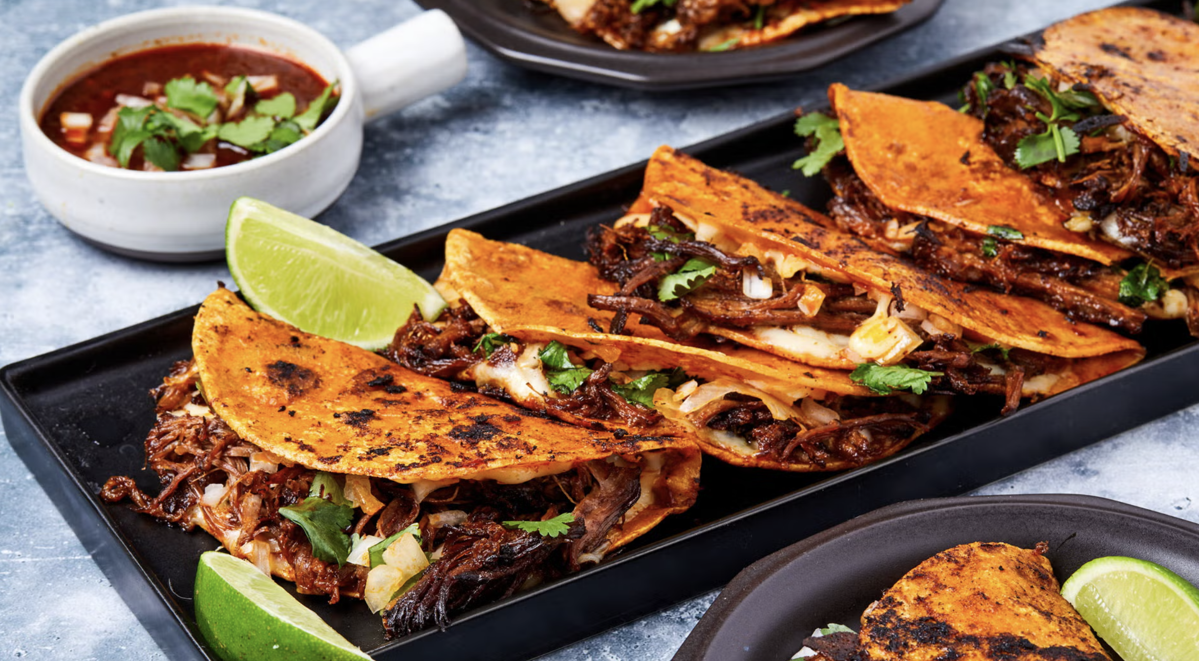 Close-up of several tacos filled with shredded beef, garnished with cilantro, served on a black rectangular platter with lime wedges, accompanied by a bowl of spicy salsa and a grilled corn tortilla on a dark plate.