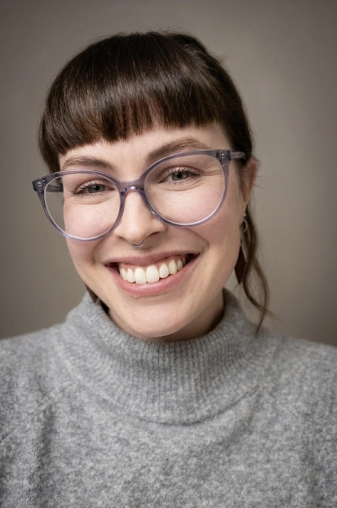 A smiling woman with short brown hair, wearing glasses and a gray turtleneck sweater against a plain background.
