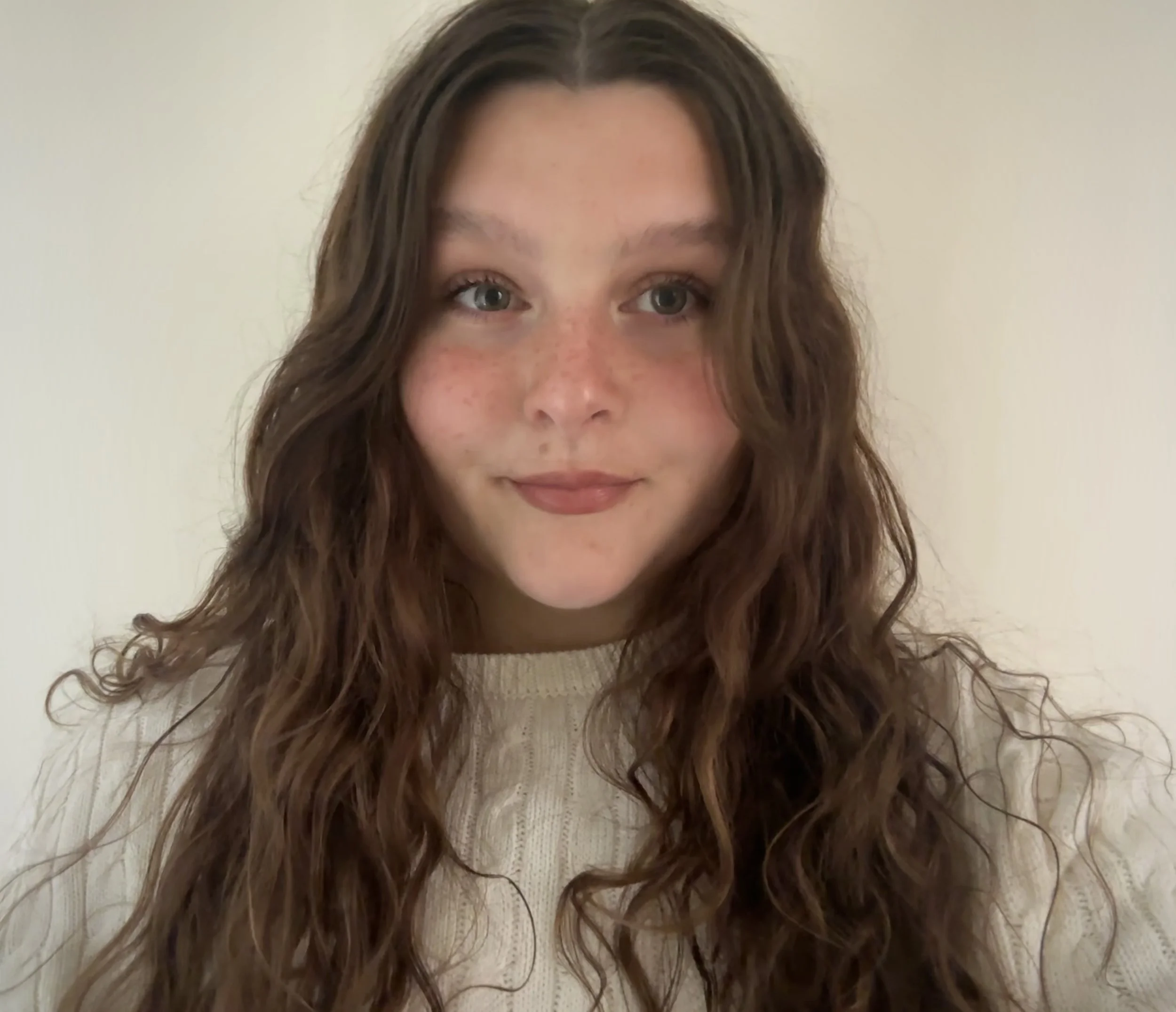 A young woman with long, wavy brown hair, blue eyes, and freckles, wearing a cream-colored sweater, looking at the camera with a slight smile.