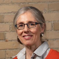 A woman with gray hair, glasses, and earrings, smiling, standing in front of a brick wall.