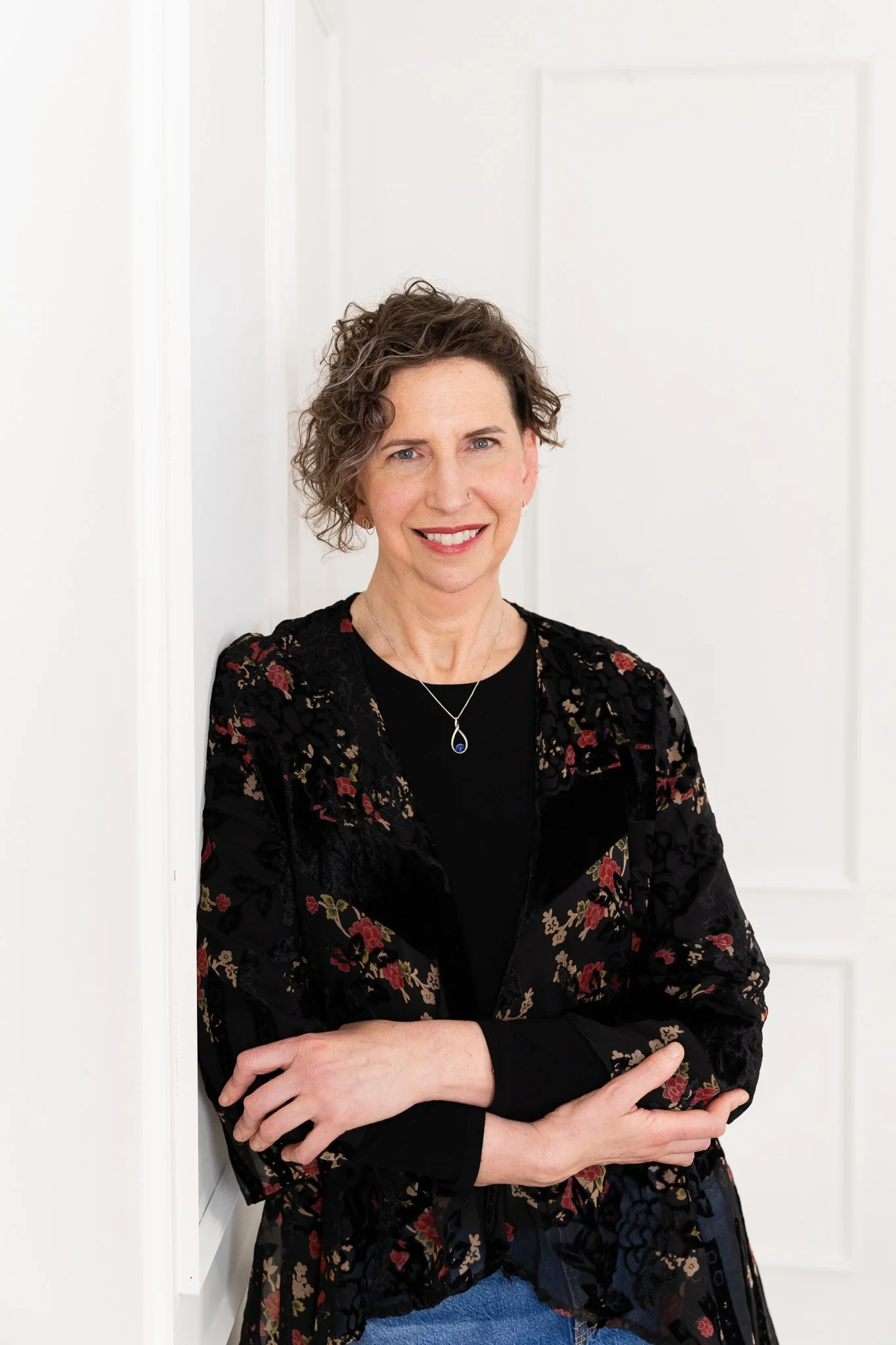 A woman with short, curly brown hair wearing a black floral jacket and black top, standing against a white wall, smiling at the camera.
