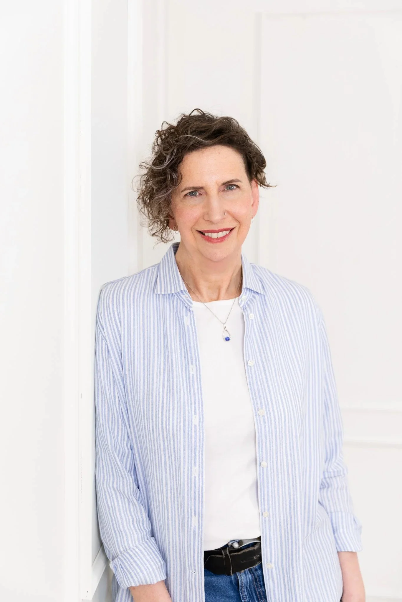 A woman with short, curly gray hair and a friendly smile, wearing a light blue striped shirt over a white top, standing against a white wall.