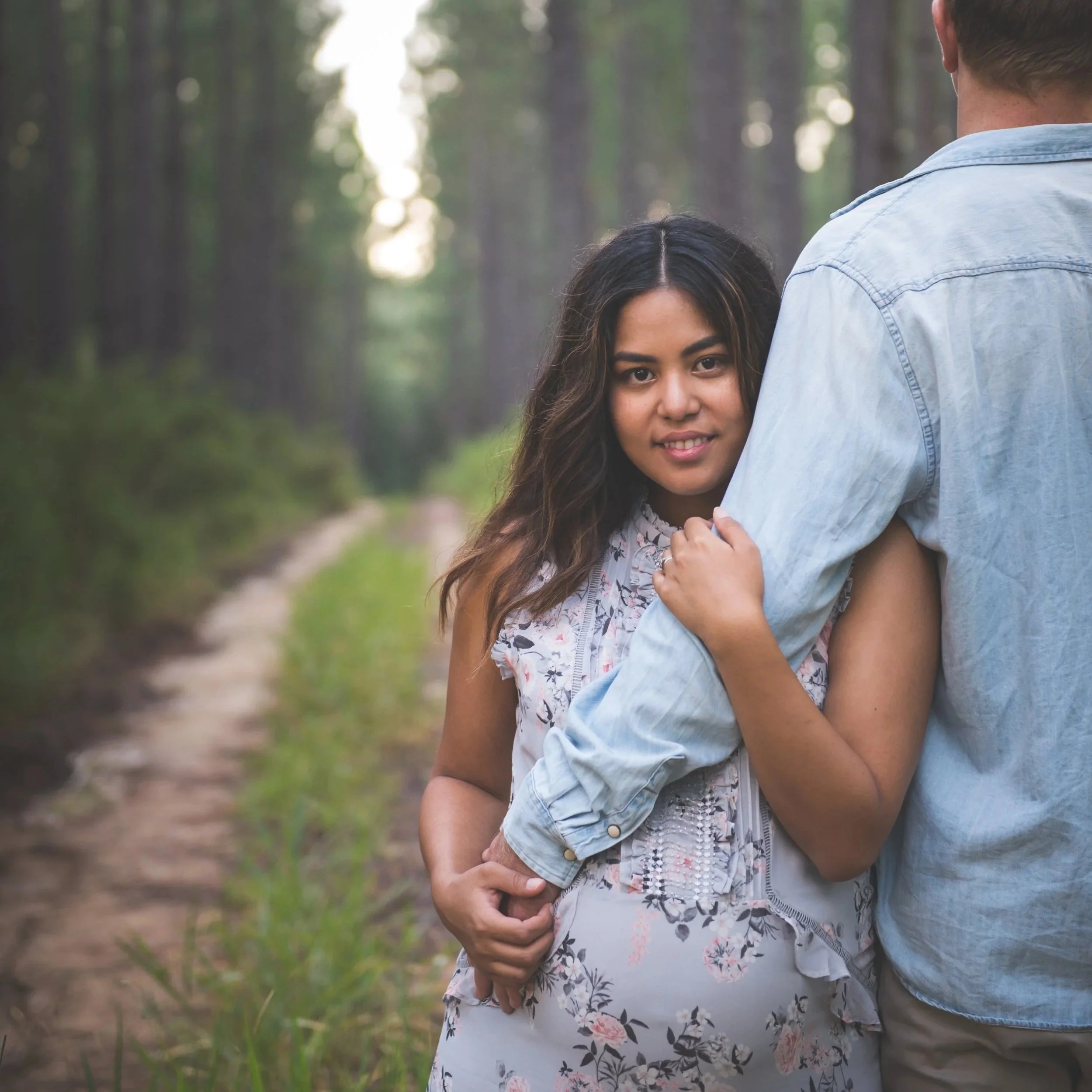 A couple hugging outdoors in a forested area, with the woman's face visible and a man standing with his back to the camera.