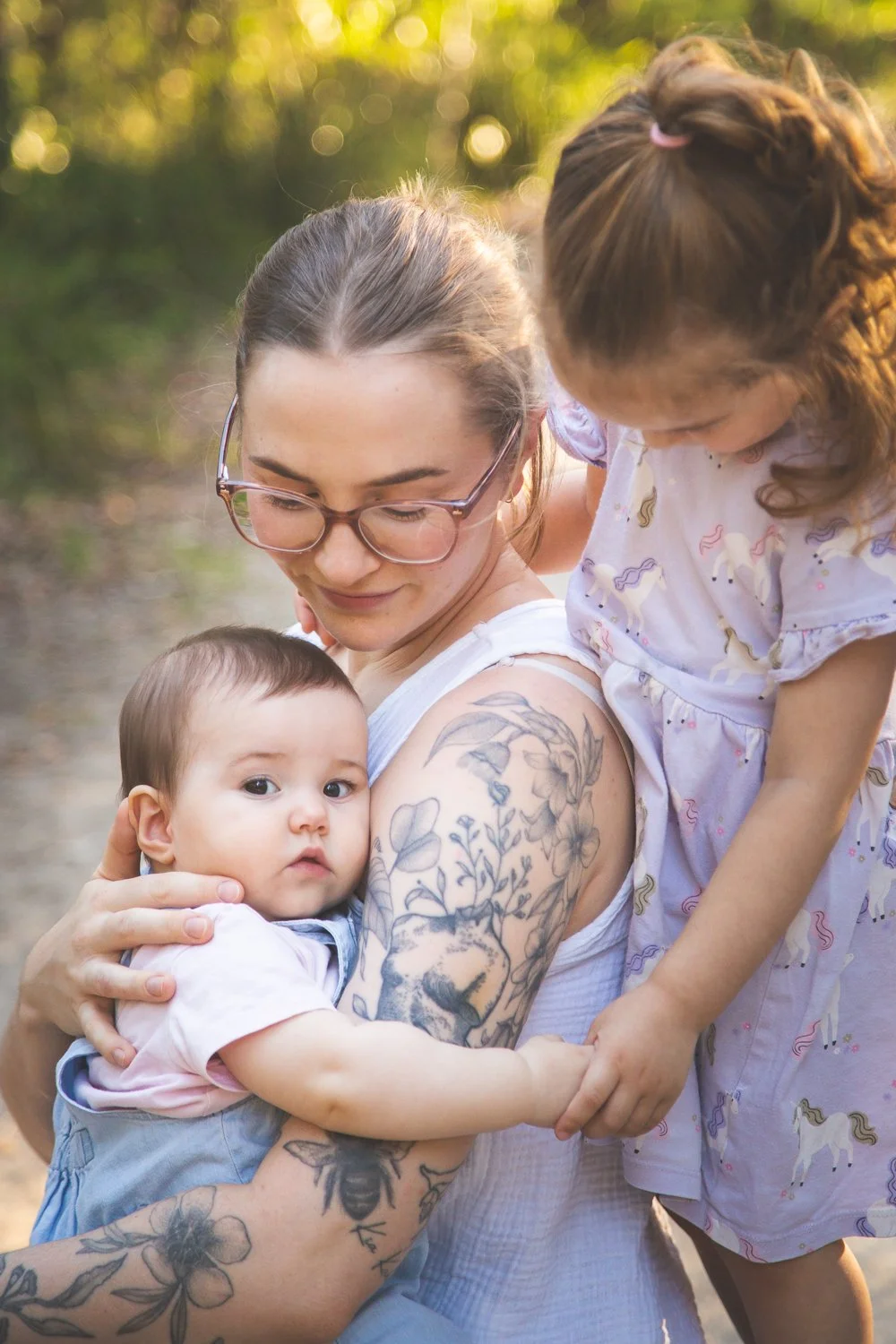 A woman with glasses and tattoos on her arm holding a baby with a serious expression, while a young girl leans over her shoulder, holding the woman's arm in an outdoor setting.