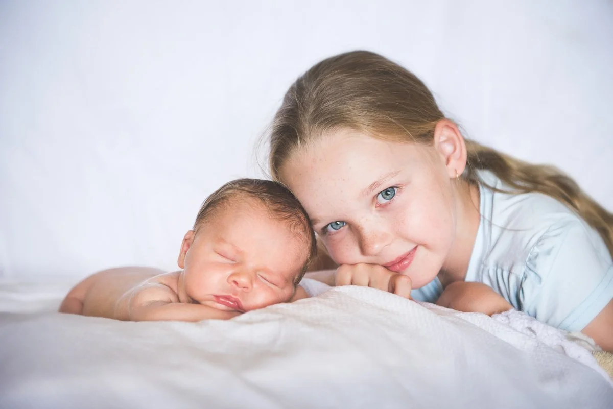 A young girl with long blond hair and blue eyes lying on her stomach next to a sleeping newborn baby with closed eyes on a soft white surface.