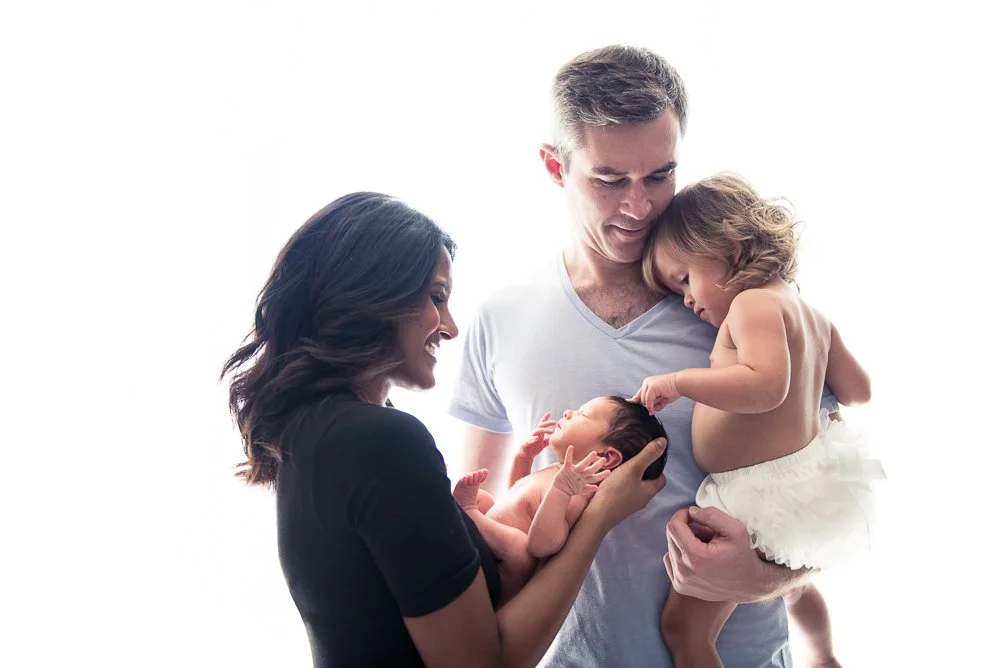Family of four, with a woman and man holding two young children, one girl and one baby, smiling and looking at each other against a white background.