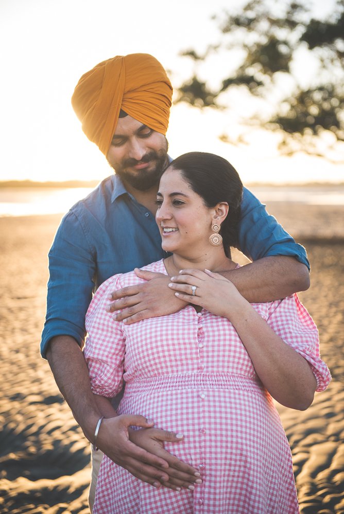 A pregnant Indian woman in a pink checkered dress and earrings, being embraced by an Indian man in a blue shirt and a yellow turban at the beach during sunset.