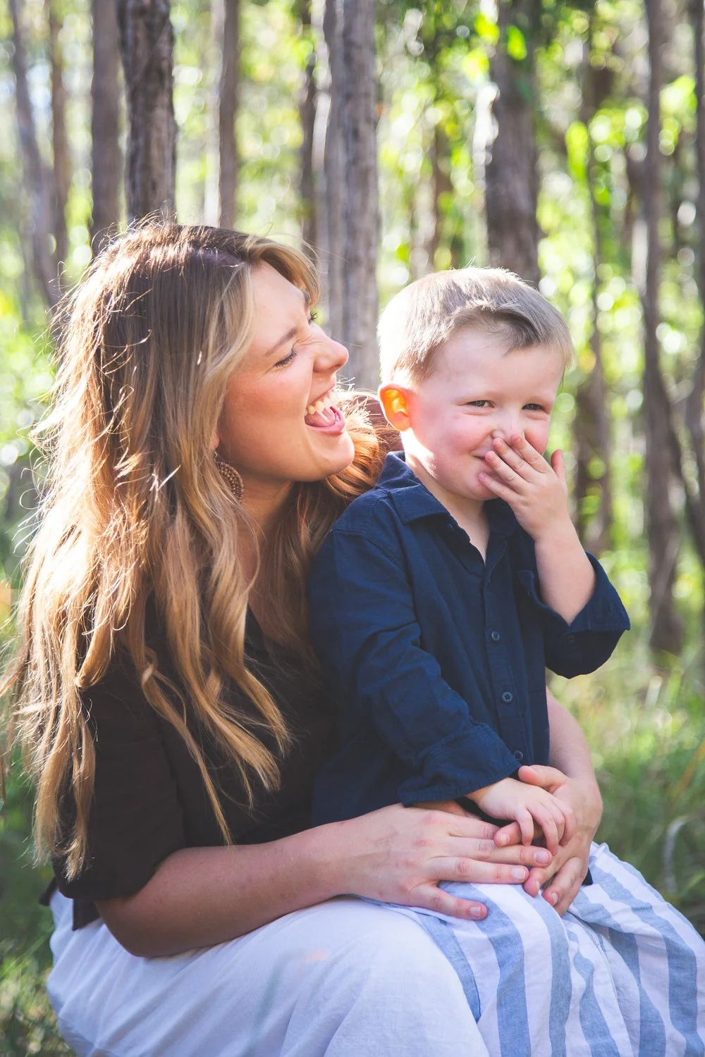 A woman and a young boy sitting in a forest with trees in the background, the woman laughing and the boy covering his mouth while smiling.