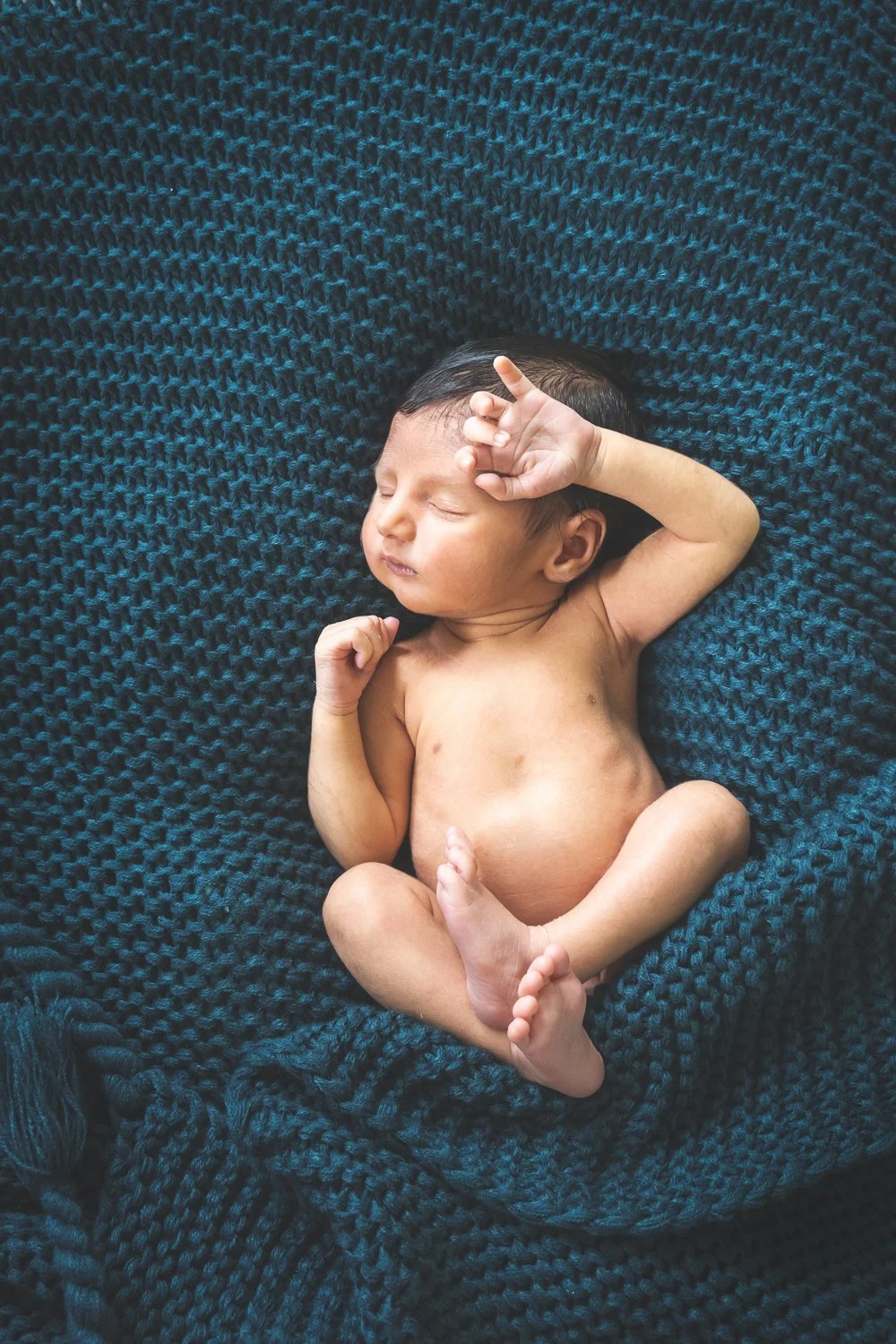 A newborn baby sleeping on a dark blue knitted blanket with a relaxed pose.
