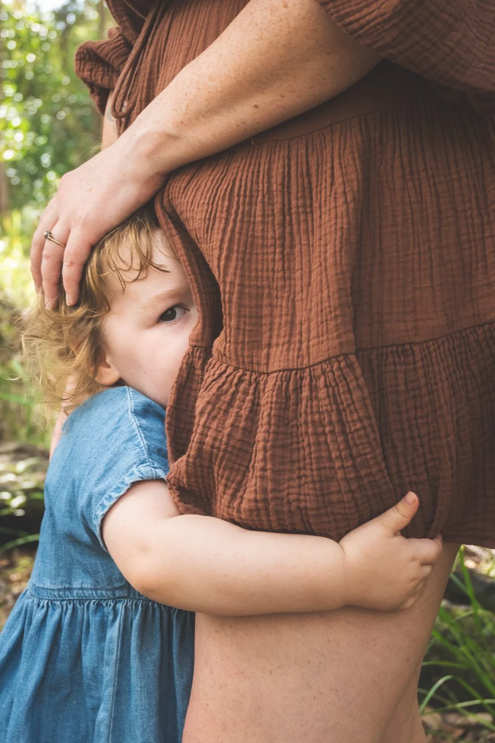 A young girl with curly red hair hugging an adult woman, who is wearing a brown, textured dress, outdoors in a natural setting with green foliage in the background. The girl is peeking out and holding onto the woman's leg.