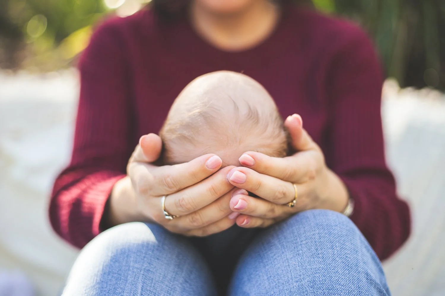 Adult holding a baby's head with both hands outdoors. The baby has light hair and the adult is wearing a maroon sweater and blue jeans.