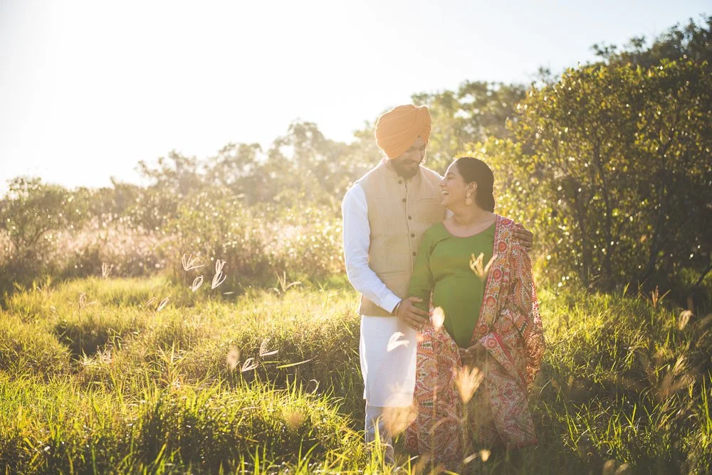 A pregnant woman in a green top and traditional Indian attire stands with her husband in a rural field during sunset.