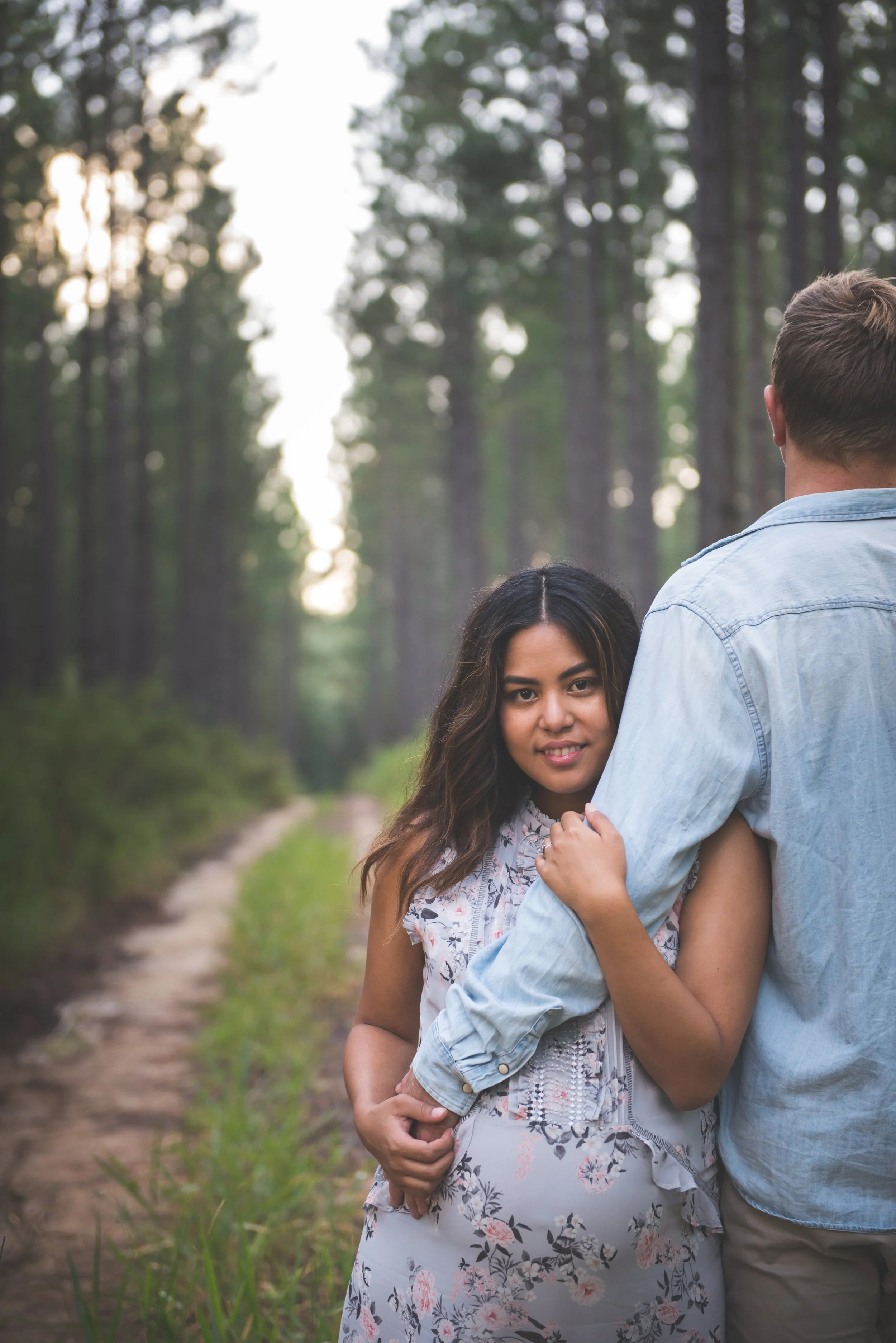 A young woman in a floral dress embraces a man in a denim shirt on a forest trail, with tall trees and a soft light background.