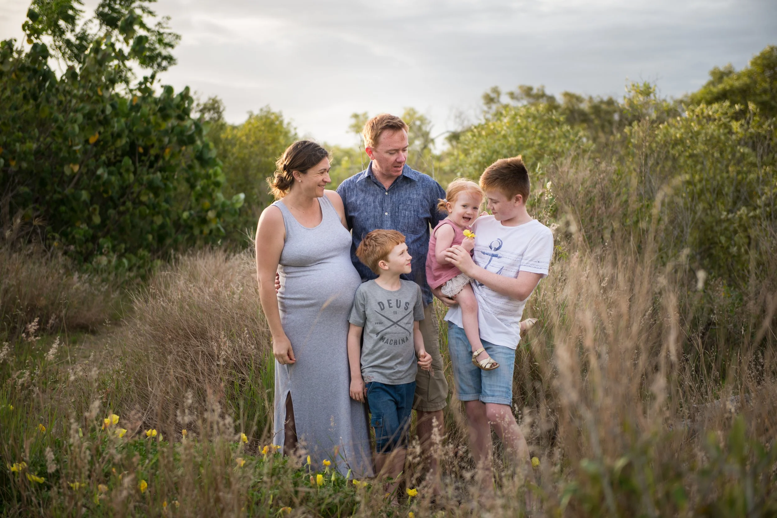 Family of six standing in a grassy outdoor field, with trees and cloudy sky in the background, smiling and spending time together.