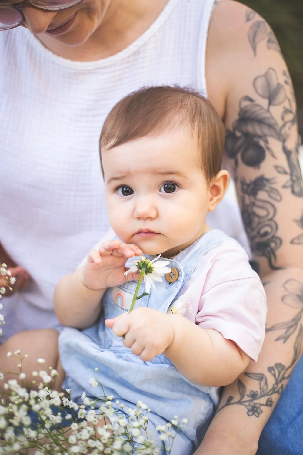 A young child with brown hair and large eyes sitting on an adult's lap, holding a white daisy flower, surrounded by small white flowers, and wearing a light pink shirt and light blue overalls.