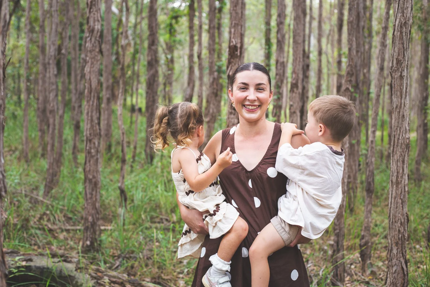 A woman smiling and holding two children in a forest with tall trees.