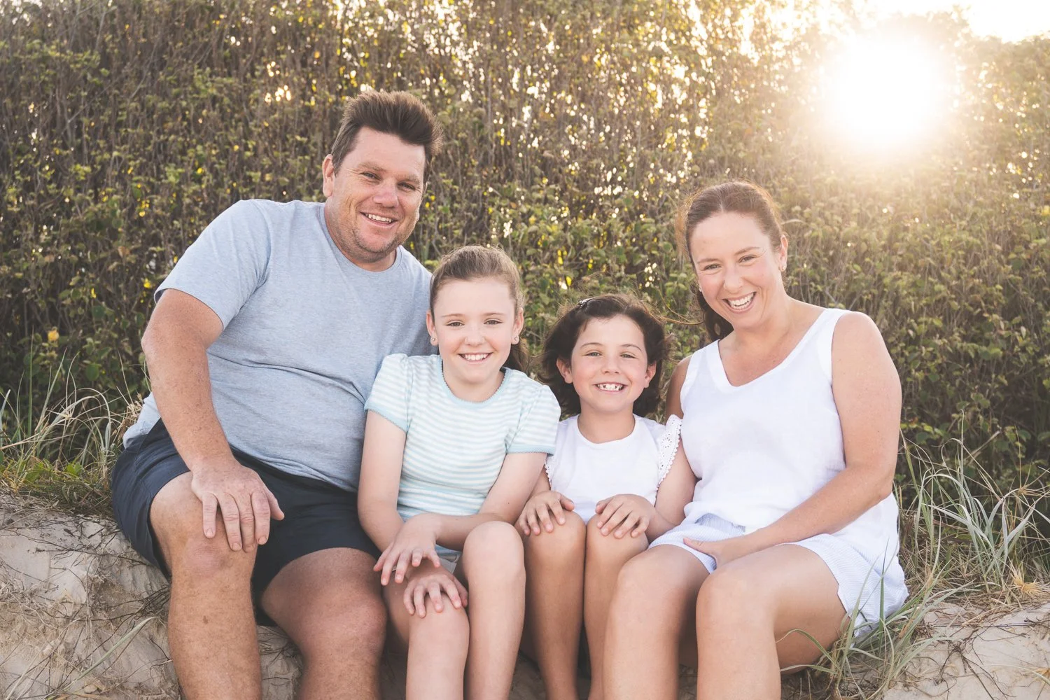 A family of four sitting together outdoors at the beach on a sunny day, with two adults and two children, smiling and enjoying the sunlight.