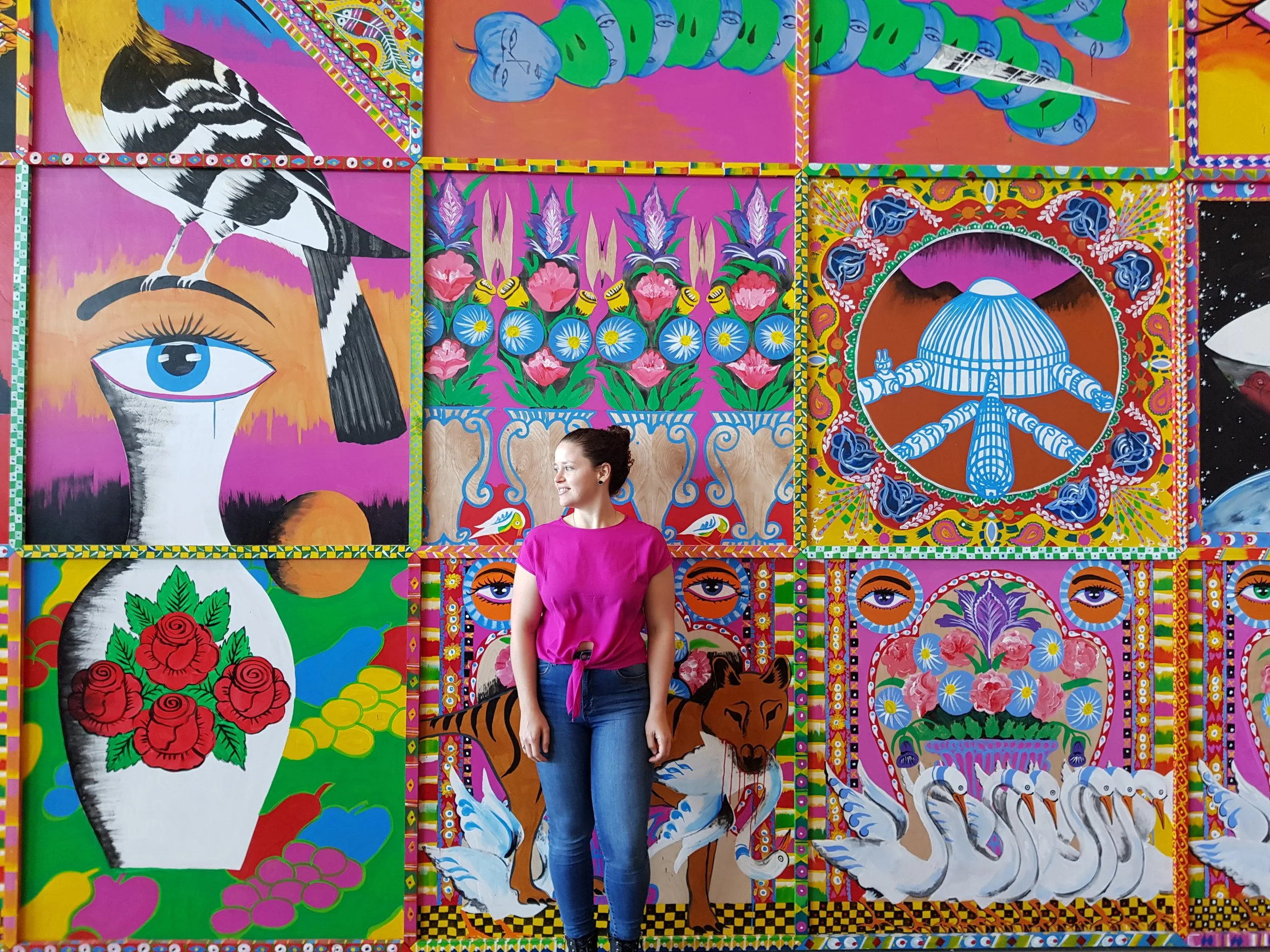 A woman in a pink top and jeans stands in front of a colorful mural featuring various vibrant, stylized images and patterns including animals, flowers, and eyes.