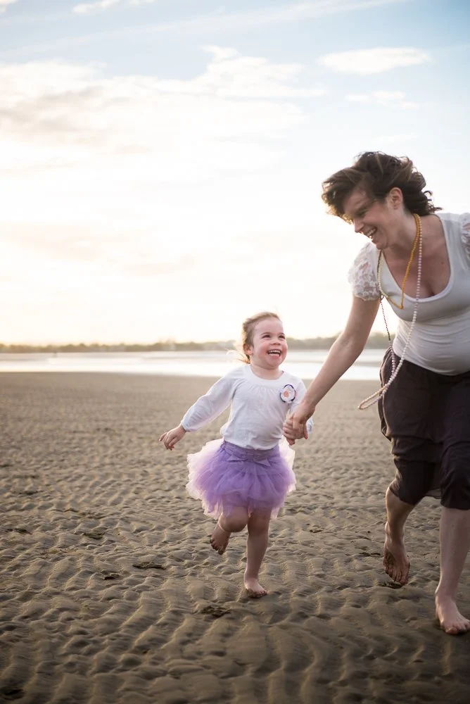 A woman and a young girl holding hands and running on a sandy beach at sunset, both smiling and enjoying the moment.