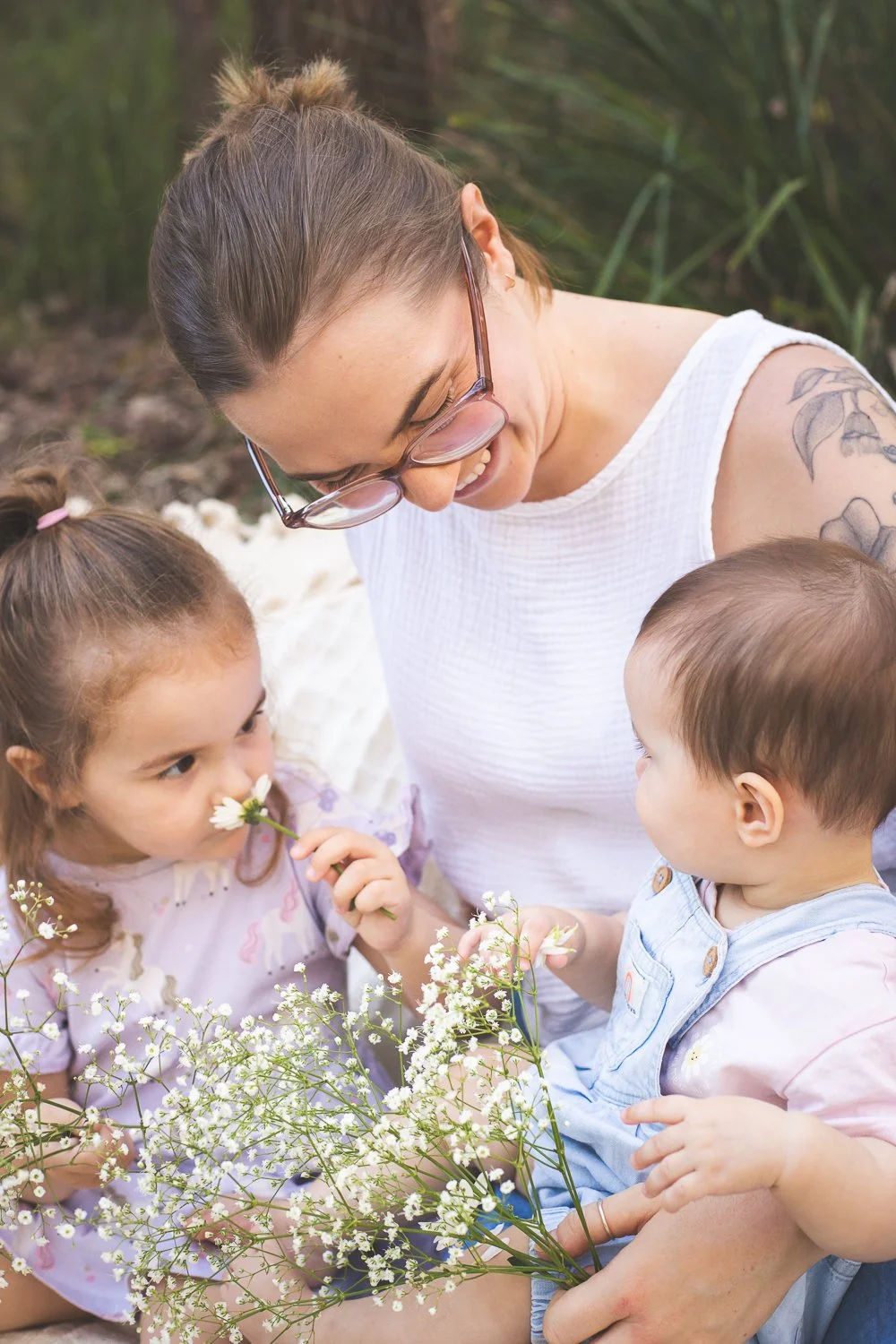 A woman wearing glasses and a white shirt smiling at a young girl and boy outside surrounded by greenery and flowers.