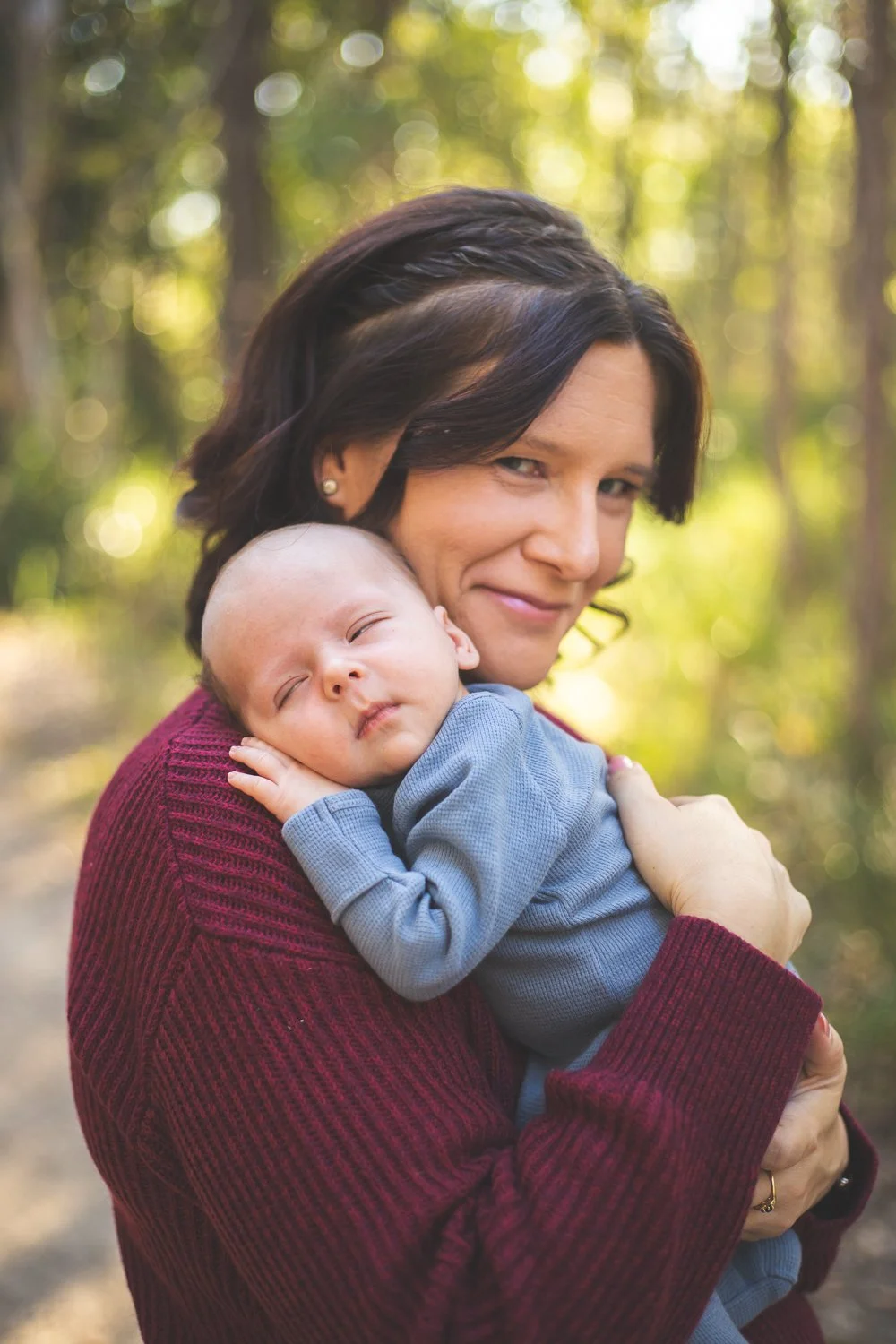 A woman with dark hair and a maroon sweater holding a sleeping baby in a blue outfit in a forest.