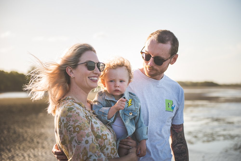 A family of three enjoying a day outdoors near a beach or lakeside with clear skies. The woman is smiling, wearing sunglasses and a floral top. The man is wearing sunglasses and a T-shirt with a patch, holding a young child in a denim jacket with a l