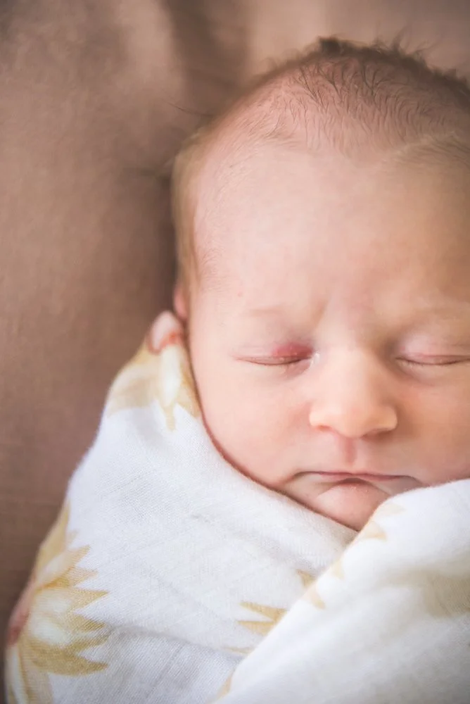 Close-up of a sleeping newborn baby wrapped in a white blanket with yellow pattern, resting on an adult's shoulder.