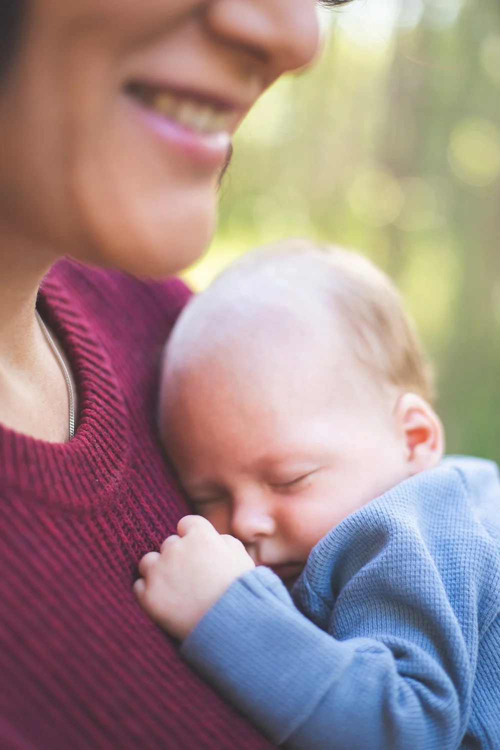 A smiling woman in a maroon sweater holding a sleeping baby who rests on her shoulder outdoors.