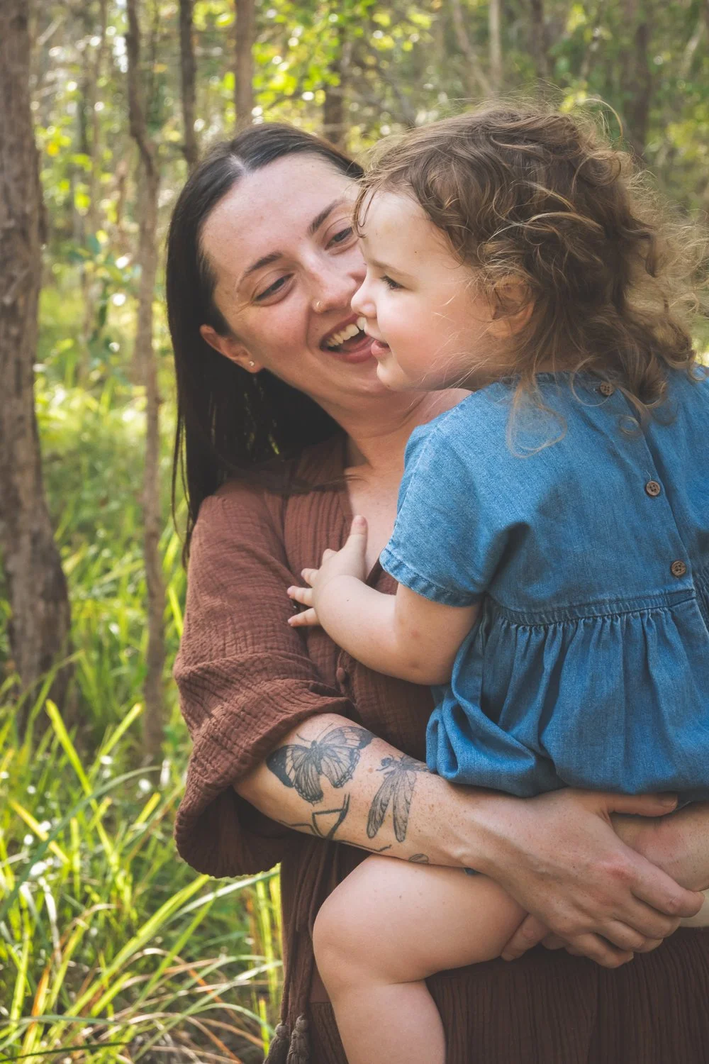 A woman holding a young girl outdoors in a wooded area, smiling and sharing a joyful moment.