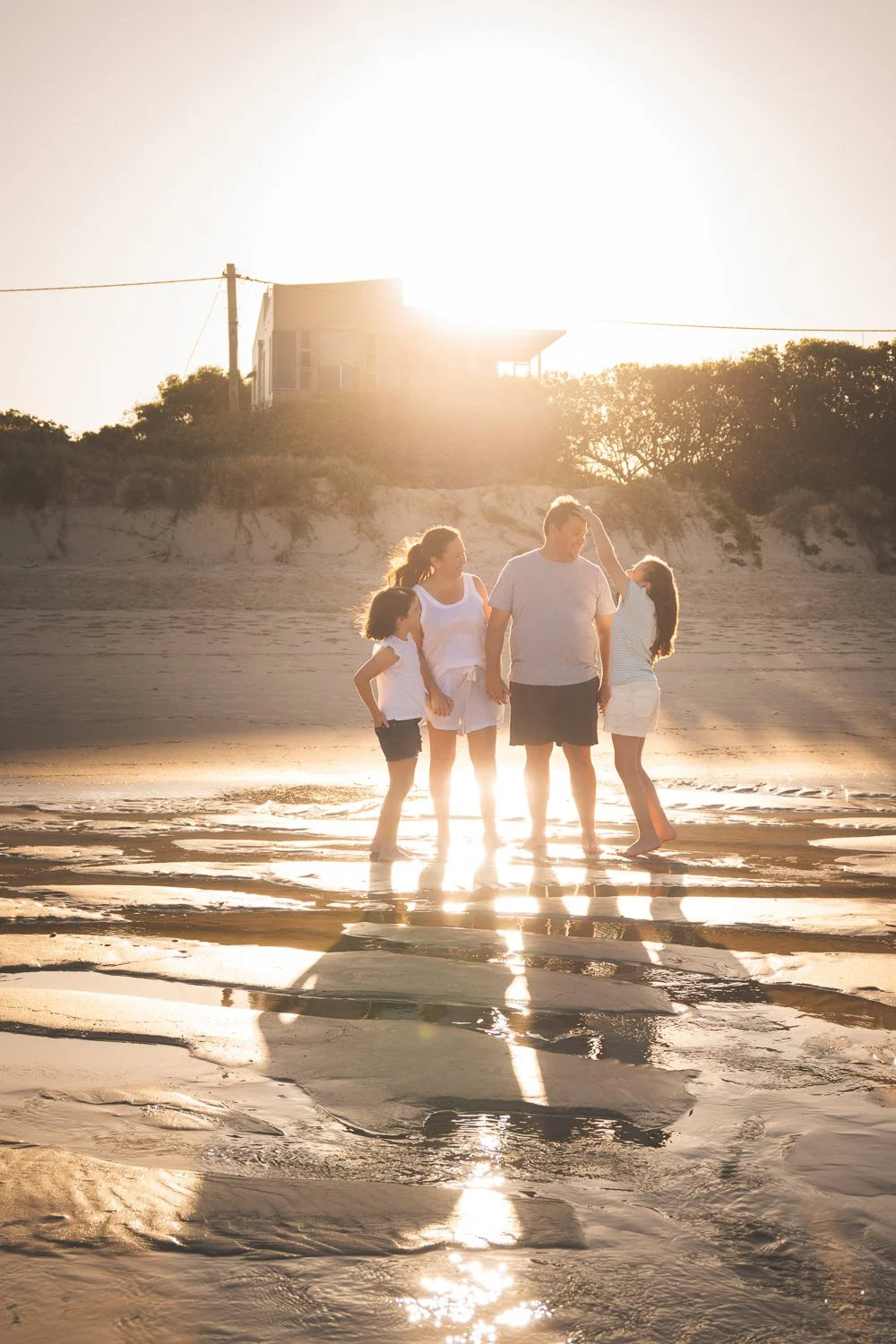 A family of four standing on a beach at sunset, with a house in the background and the sun shining brightly behind them.